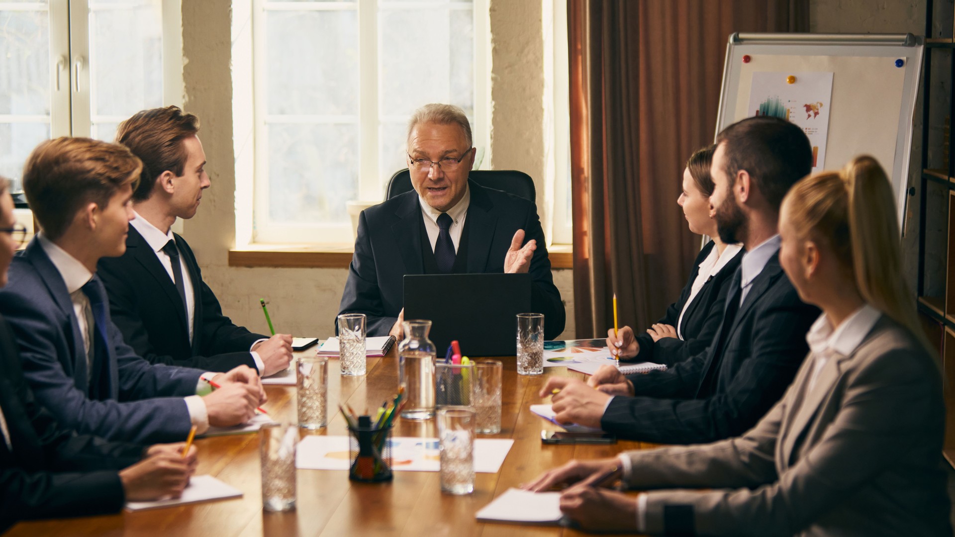 El líder empresarial senior realiza una presentación a un grupo de jóvenes profesionales durante la reunión del equipo, con gráficos y tablas visibles en el fondo.